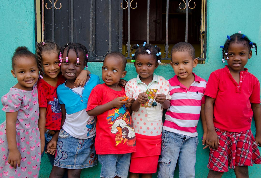 Seven children stand together. Some hold hands while other have their arms around each other's shoulders. They stand infront of a seafoam green wall.