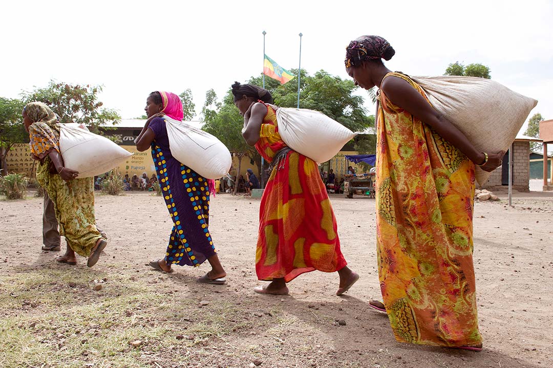 Ethiopian women wearing colourful, full length dresses, carry bags of rice across a dirt square.