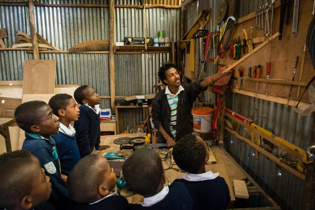 A young teacher uses a long piece of wood to point to different tools hanging on the wall. A number of carpentry students look on.