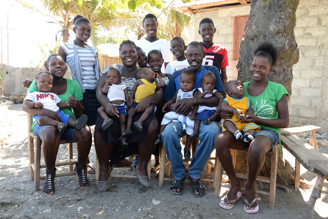 A husband and wife sit on chairs in the shade of a tree. Gathered around them are their thirteen children, including baby quadruplets.