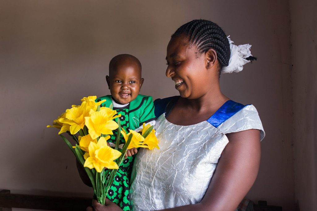 Esta, a Ugandan mother, holds here infant daughter, Zena and bouquet of yellow flowers. Both smile.