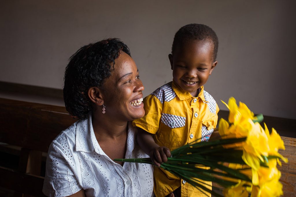 Lucy, a Ugandan mother holds her infant son, Crispin and yellow bouquet of flowers. They are smiling at eachother.