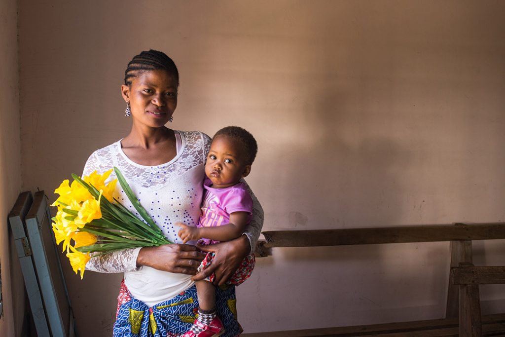 Neema, a Ugandan mother, stands inside a building holding her daughter, Tumaina and bouquet of yellow flowers.
