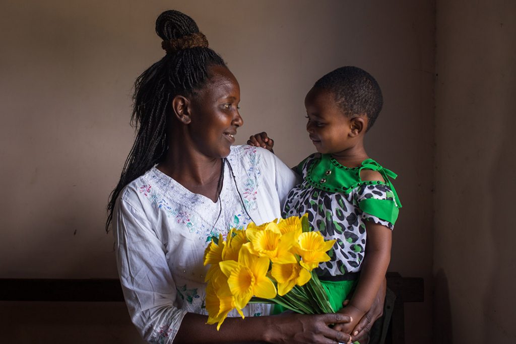 Winner, a Ugandan mother holds her infant son, Godnester and yellow bouquet of flowers. They are smiling at each other.