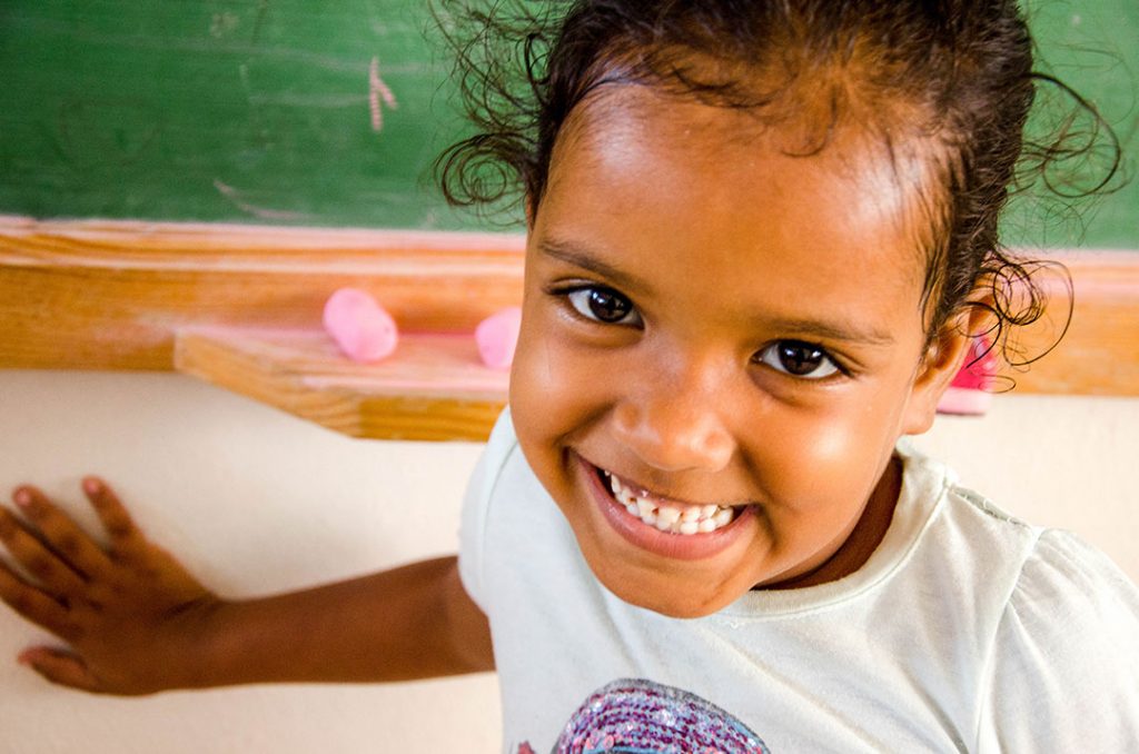 Close-up of a young female child, little girl smiling. She is wearing a white shirt with her hand on the wall. A green chalkboard and chalk is in the background.