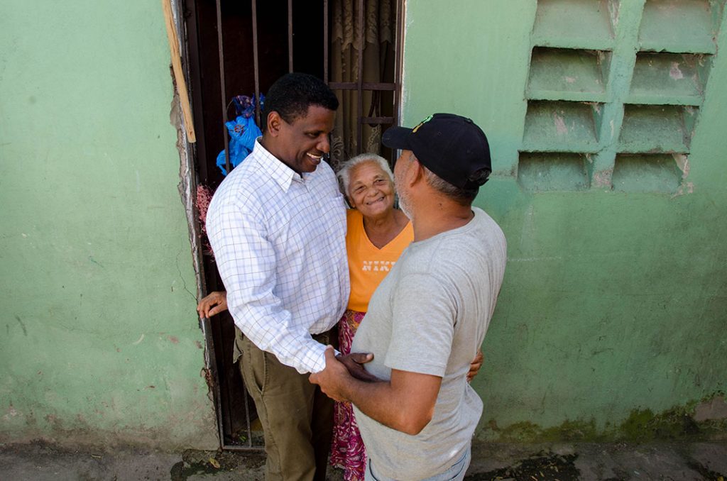 Pablos stands and talks with a man and an older women. They stand in the door way of a vibrant, green house.