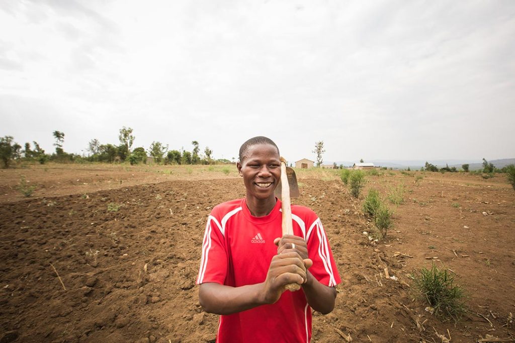 A man stands in a freshly tilled field, a garden hoe resting on his shoulder.