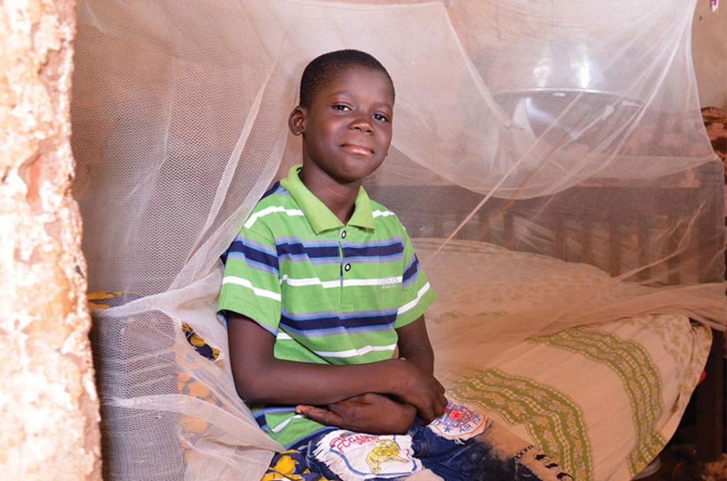 A boy sits on his bed. A mosquito net is draped behind him.