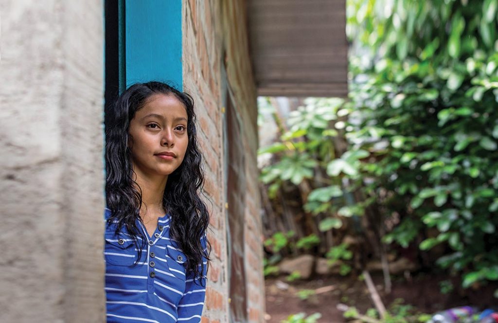 A teenage girl stands in a doorway, looking off into the distance.