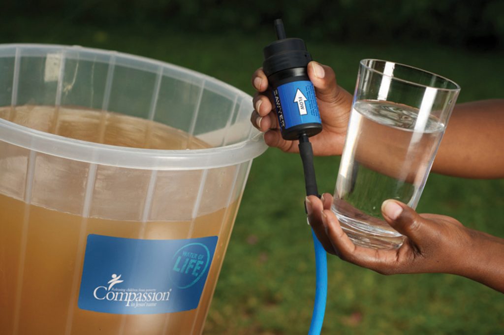 A water filter held up between dirty water and a glass of clean, filtered water.