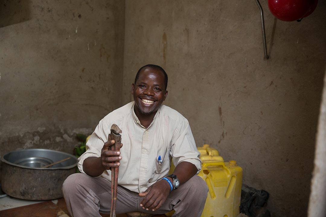 A man wearing a white shirt and holding plumbing equipment smiles brightly.