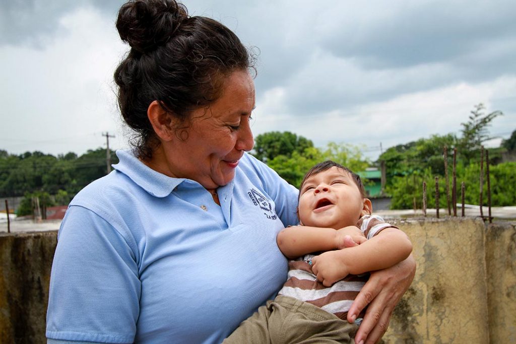 A compassion worker holds a baby boy in her hands and smiles at him.