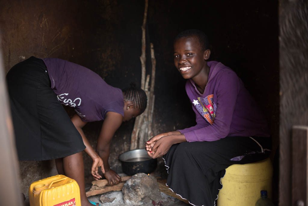 A girl sits inside her families hut as someone cooks over a fire.
