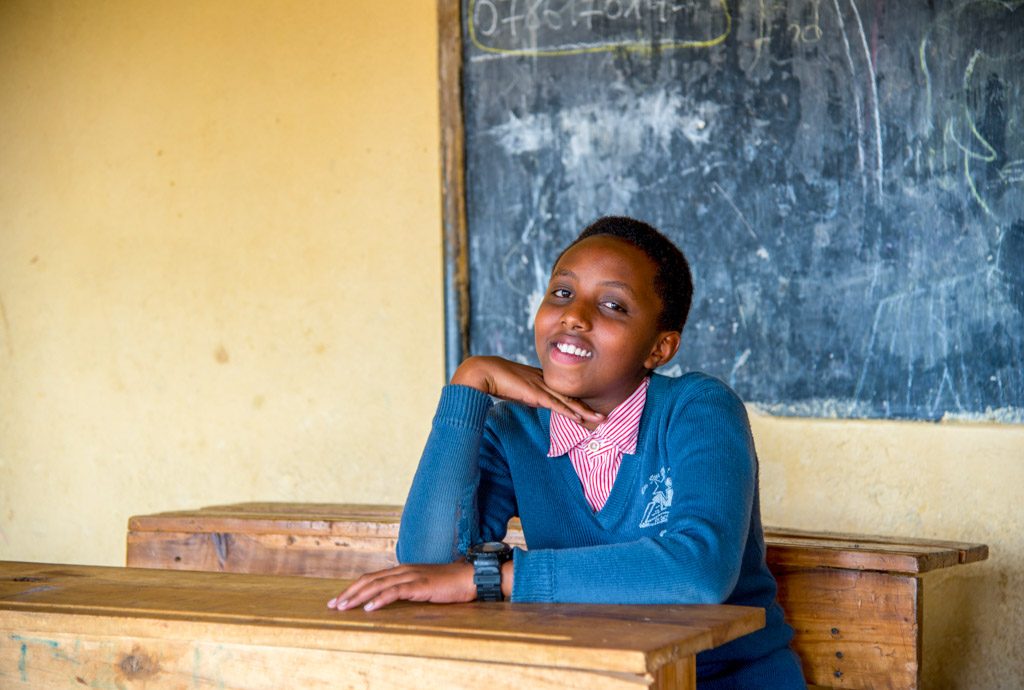 A girl sits at wooden school desk with a chalk board in the background. She leans with her chin on hand her elbow on the desk.
