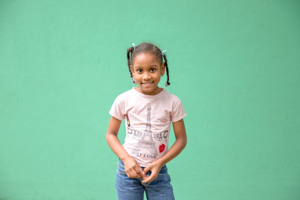 A young girl stands in front of an emerald green wall. She wears a pink t-shirt with the Eiffel tower on it, she wears jeans.