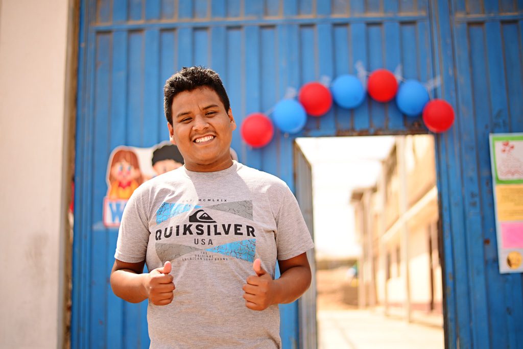 A young man stands in front of a blue wall with balloons on it. He wear a gray, quicksilver shirt and give two thumbs up.