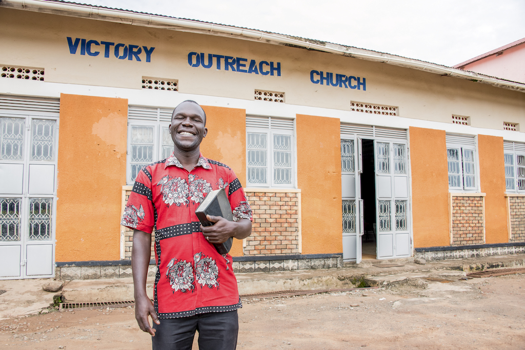 A man stands holding a Bible in front of a building that says "Victory Outreach Church".