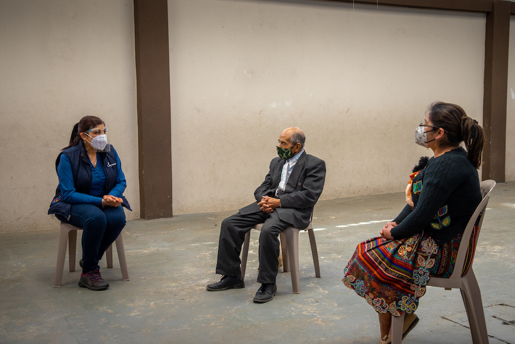 Partnership Facilitator Virginia sits with a pastor and Compassion centre director. They are social distancing and wearing protective face masks.