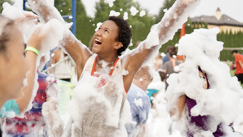 A teen participates in a game at camp. She is smiling with her arms in the air.