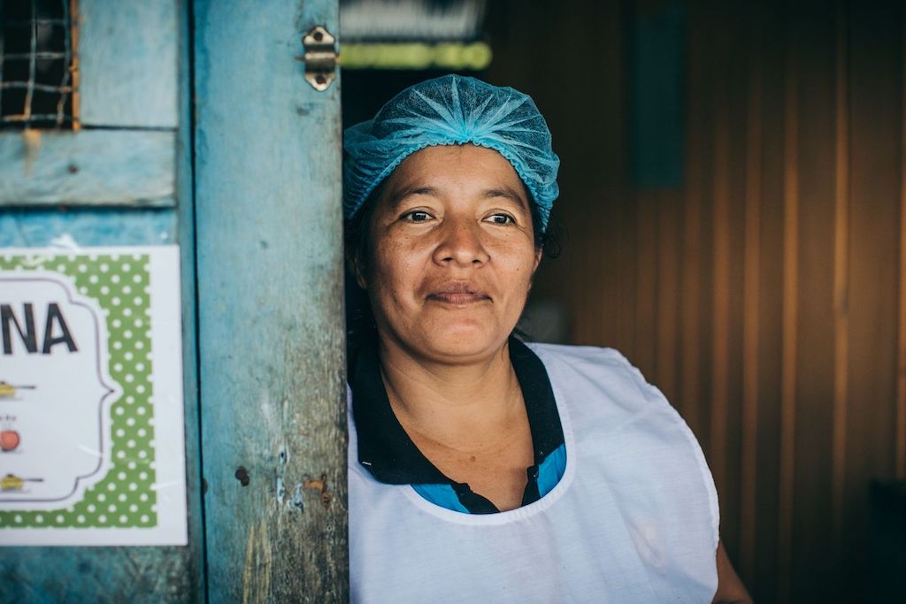 A woman in a white smock, apron and blue hair net leans against a blue door in a doorway.