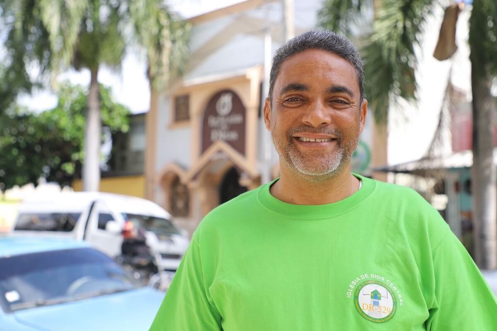 Asunción, a man in a green shirt stands next to the street. There are cars, tropical trees and a church in the background.