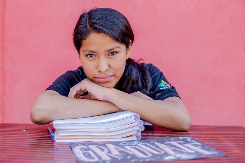 A girl leans against a stack of workbooks on a table, with her arms crossed.