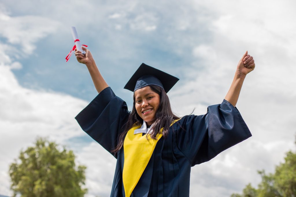 A girl in a graduation gown and cap raises her hands in celebration, holding a diploma.