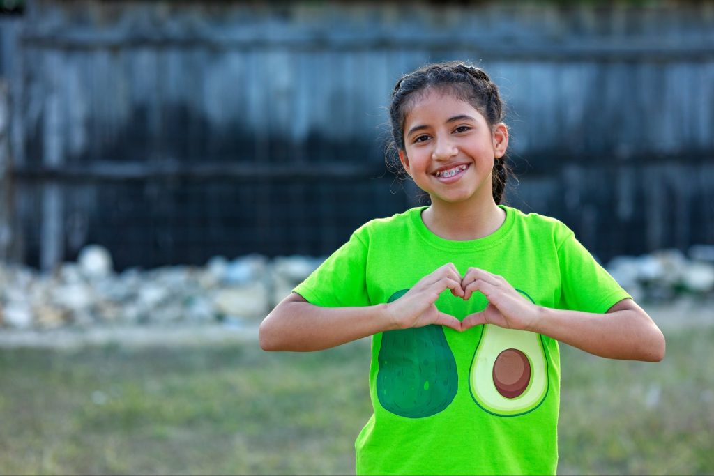 A young child smiles while making a heart with her hands.