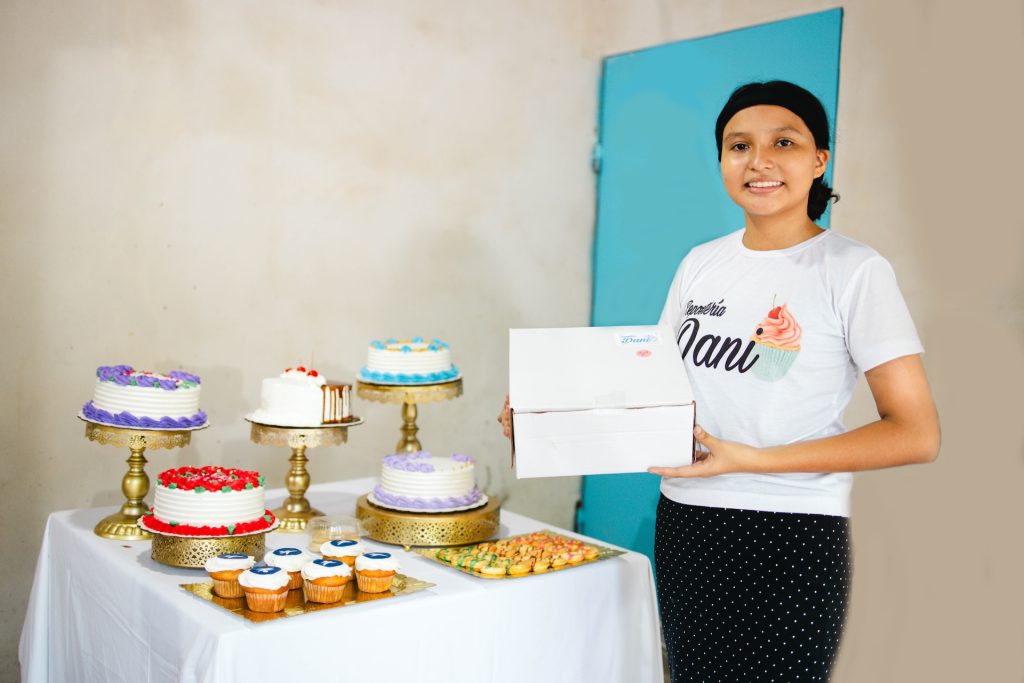 Daniela posing in front of many cakes baked and decorated by her. She is holding a box with a cake to be delivered. She is smiling.