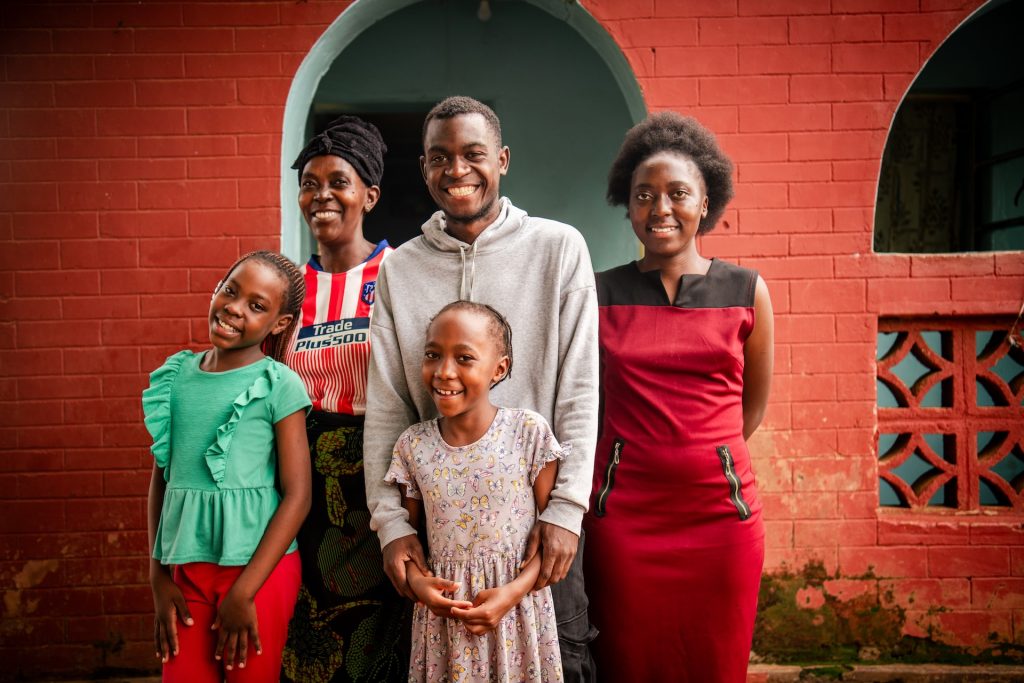 Joseph and his family smile just right outside their house. Joseph stands in the middle of his family with his younger sister standing in front of him, his arms cupped around her arms. He is wearing a gray hoodie, and his sister is wearing a gray dress with a butterfly pattern. His mom and older sister are on either side of him, and another younger sister stands in front of his mom. Behind them is a brick building painted red with an arched doorway and arched window behind them.
