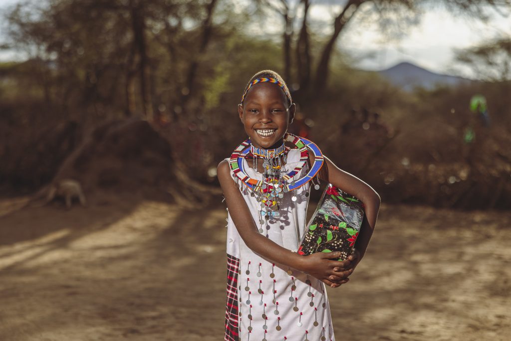 A young girl from Kenya wearing traditional clothing and jewelry holds a Christmas present and smiles.