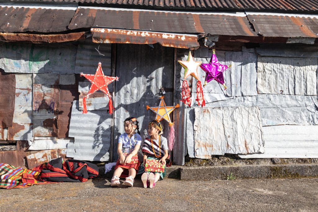 Two children from the Philippines sit on their front stoop of a tin home decorated with colourful Christmas stars.