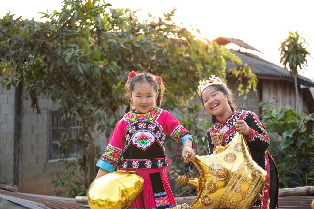 Two girls from Thailand wear traditional outfits and hold gold star balloons while laughing.