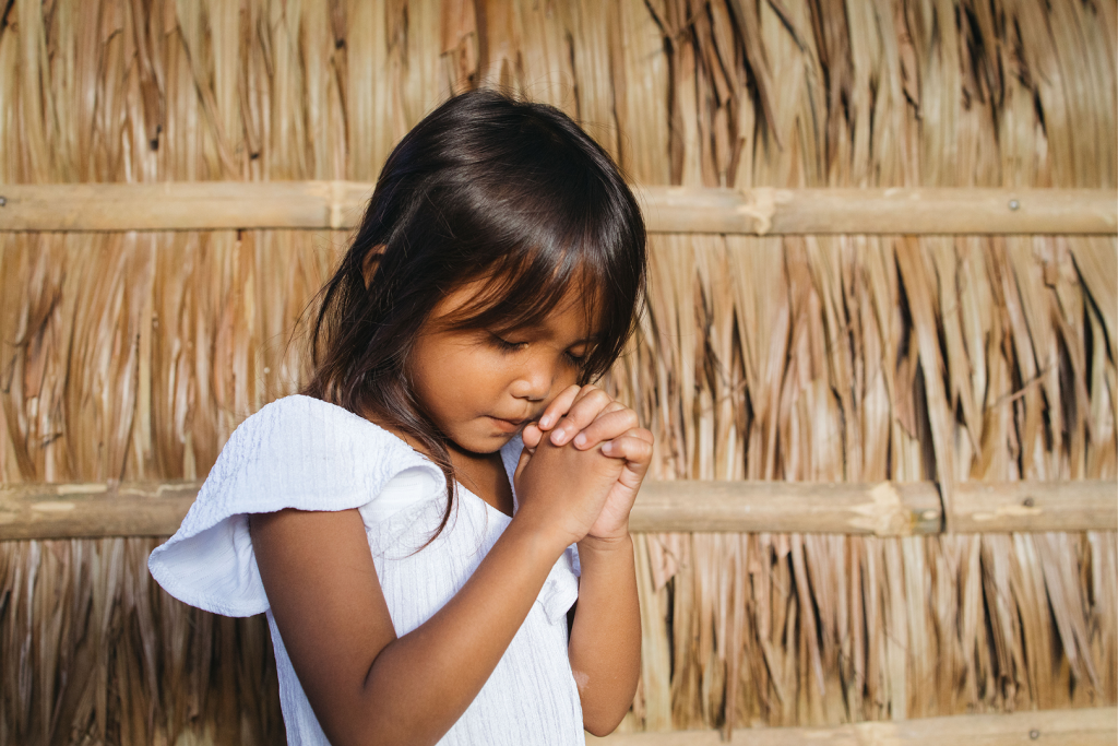 A young girl in a white shirt clasps her hands in prayer.