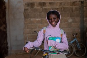 A young girl wearing a pink sweater shit and a backpack smiling