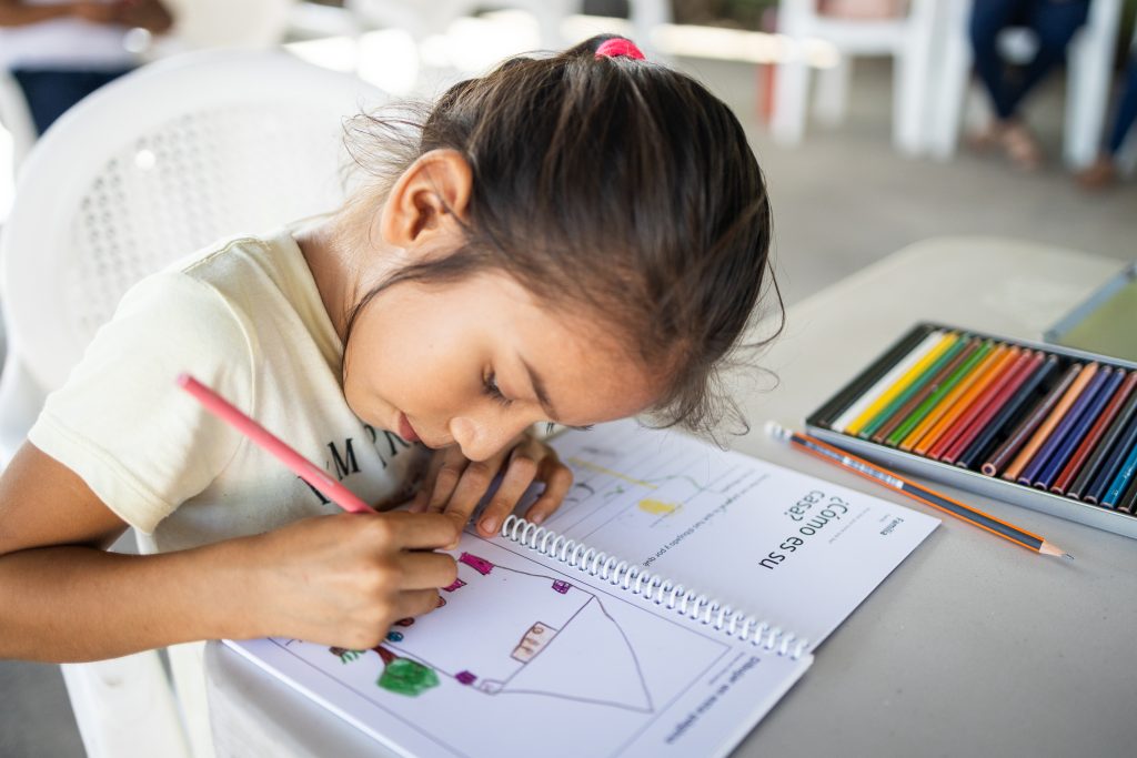 A little girl is at a desk with a set of coloured pencils drawing a picture of her neighbourhood on paper.