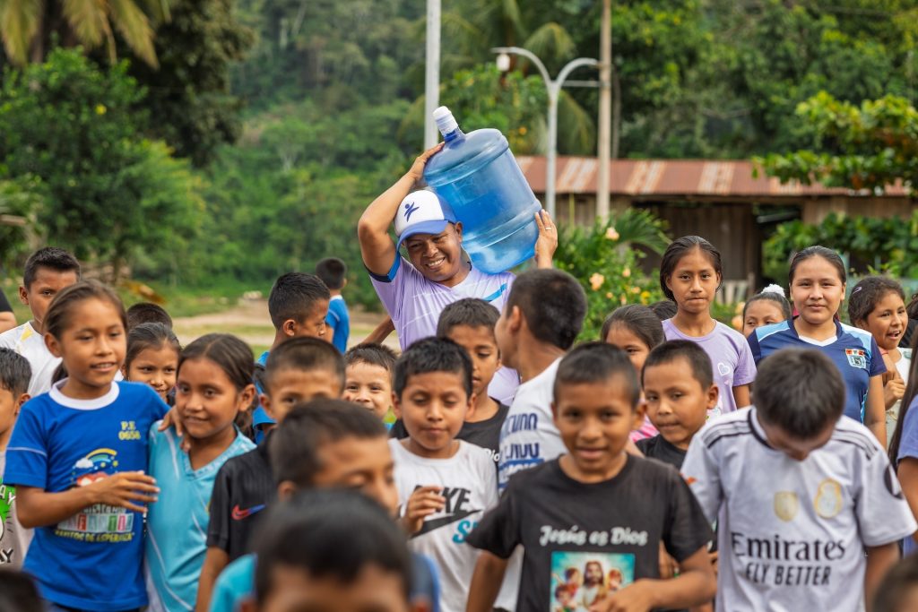 Children in Tununtunumba, Peru are receiving the centre director, who is transporting the safe water for them. The director is holding the water container on his shoulder.