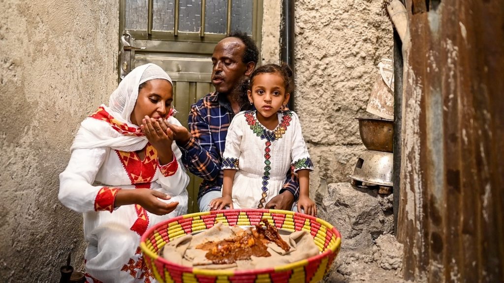 Elbetel sits with her parents, Anteneh and Tsehaynesh, at home as they celebrate Easter. Anteneh is performing 'gursha' for Tsehaynesh, or feeding one another from the hand. A bowl of food is before them in a colourful basket.