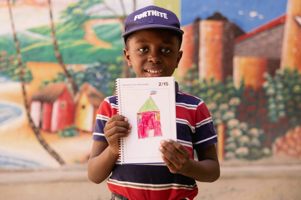 A child wearing a hat and a striped shirt smiles holding up a drawn picture of a house.