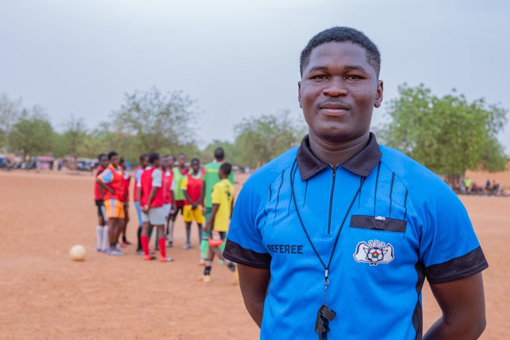 Raoul, a talented district referee, is standing at a soccer field in his community. He has his hands tucked behind him as he smiles at the camera. He is wearing a blue polo shirt with a whistle around his neck. Behind him is a soccer team huddled together.
