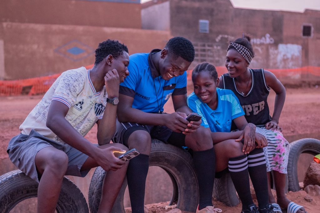 Raoul and his three referee assistants are joyfully debriefing a soccer game recorded on a phone. They sit on a row of tires next to a soccer field as they smile and Raoul is between them holding the phone in view. He is in a blue polo shirt with black shorts and the referee next to him is in the same.