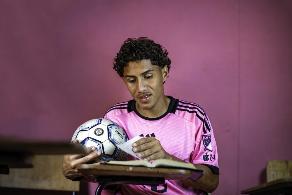 Alejandro is holding a soccer ball and a Bible as he sits at a desk inside a center classroom. He looks down at his Bible and turns a page. He is in a pink sports jersey. 