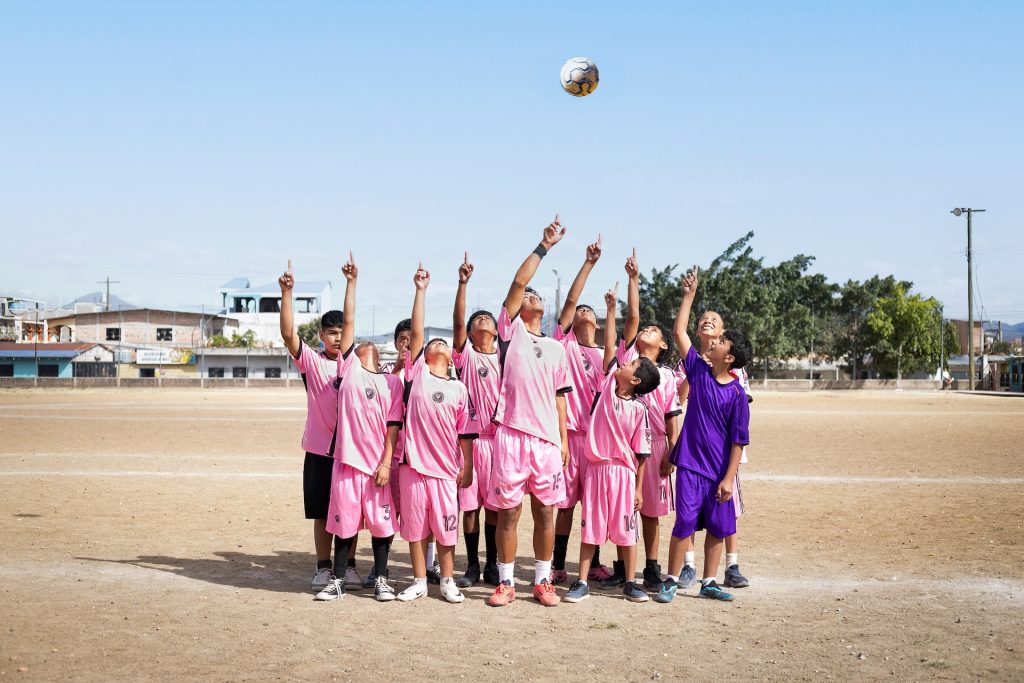 Alejandro stands in a group with the center’s younger participants during training at a local soccer field. They all point up and look to the sky as a soccer ball flies above them. Most of them are wearing matching pink soccer jerseys and pink shorts. 