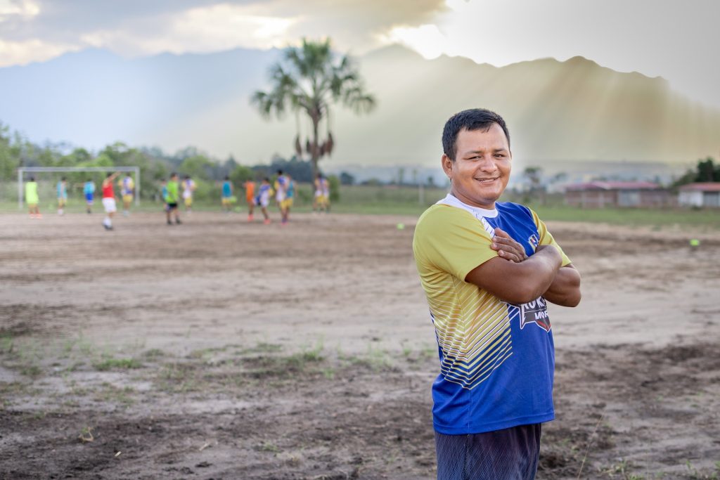 Portrait of Jose, soccer coach and centre staff, standing in the soccer field. He is wearing a blue, yellow and white top. His arms are crossed at his chest. He is smiling.
