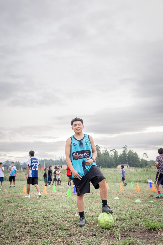 David, one of the boys at the soccer academy, smiles and poses for a photo in the soccer field. His left foot is holding the soccer ball in place. He is wearing a light blue top.
