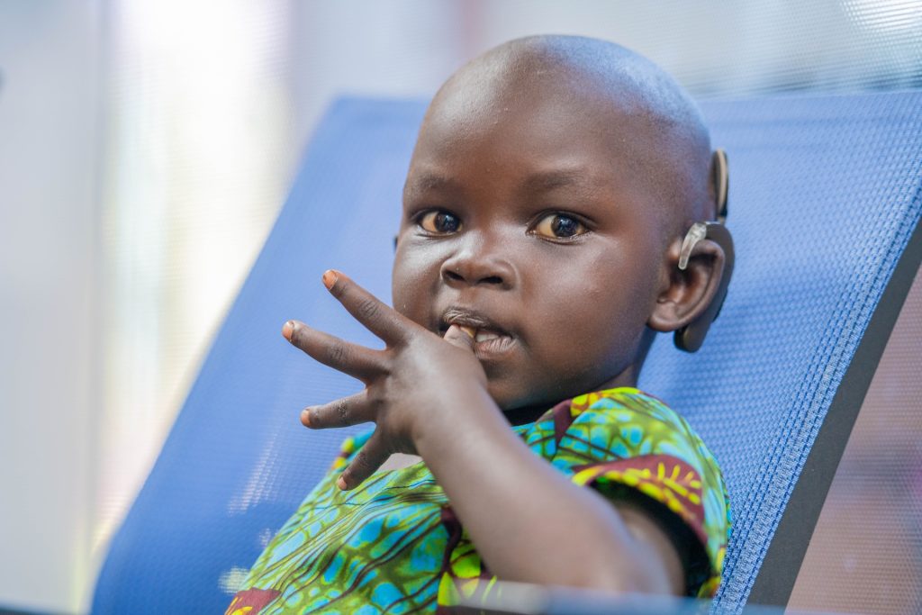 Deborah sits in a blue chair looking at the camera with her thumb in her mouth. She has implants around her left ear and is wearing a patterned shirt with blue, green and yellow tones.