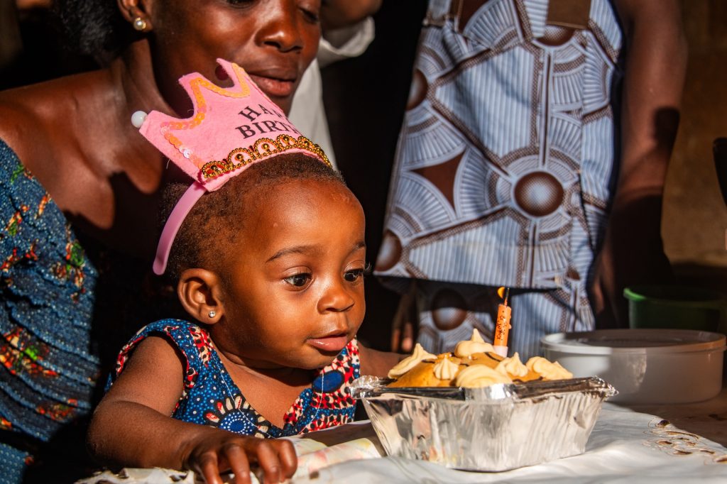 Emmanuella, in a blue dress and pink birthday crown, is sitting on her aunt Mawulawoe's lap and looking at the birthday cake on the table in front of her. The cake is in a metal pan on top of a white tablecloth and has a lit candle on top of it.