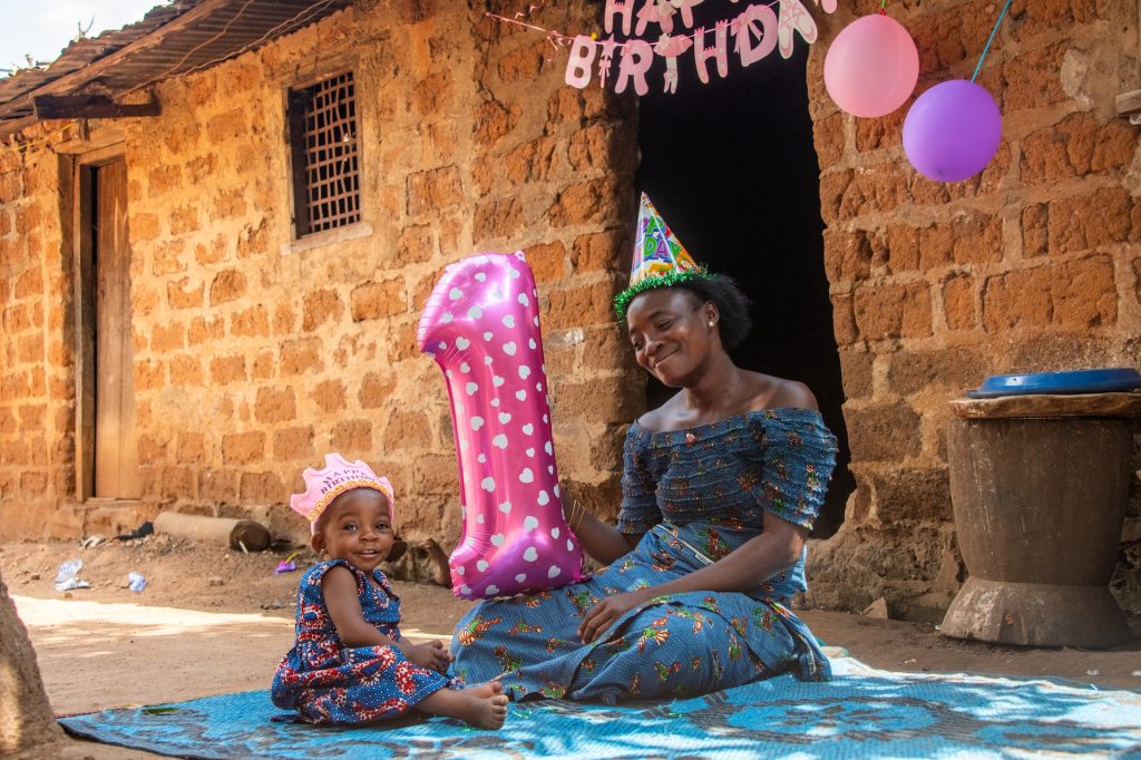 Emmanuella is sitting on the ground with her aunt Mawulawoe. The baby is wearing a pink paper crown and Mawulawoe is wearing a birthday hat. They are both wearing blue dresses. Mawulawoe is holding a big pink balloon in the shape of the number 1, and there is a banner that says “HAPPY BIRTHDAY” hanging over the doorway of the brick building next to pink and purple balloons.