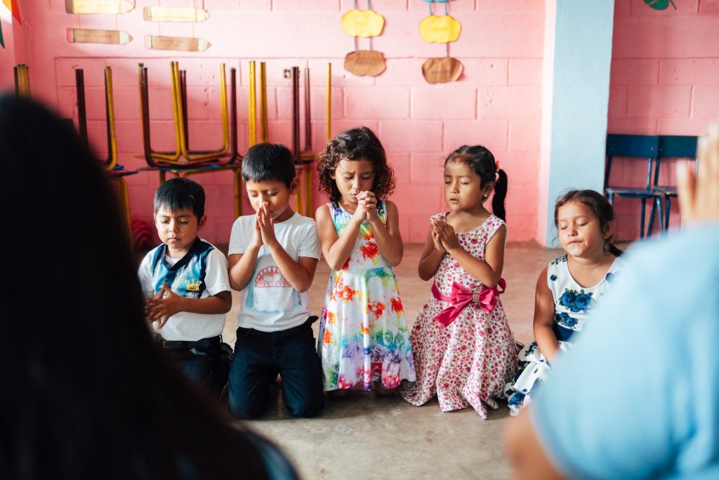 Carlos, Esdras, Angie, Marjorie, and Lissa pray on their knees in the Compassion center's classroom, in front of their tutors, as part of the Bible lesson where they are learning about the importance of prayer. Carlos and Esdras are in white t-shirts and jeans. Angie is in a white dress with a colorful pattern, Marjorie is in a white dress with a hot pink flower pattern, and Lissa is in a white dress with blue flowers. Behind them are cinderblock walls painted pink with chairs stacked up along it. 